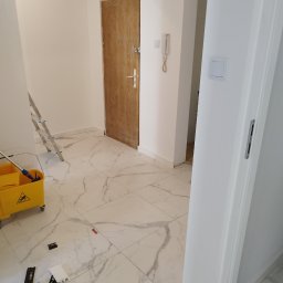 Artur Sarnecki - Wnętrze przedpokoju w trakcie malowania: białe ściany, marmurowe płytki podłogowe, żółty wózek z mopem, drabina oparta o ścianę, drzwi wejściowe w tle.