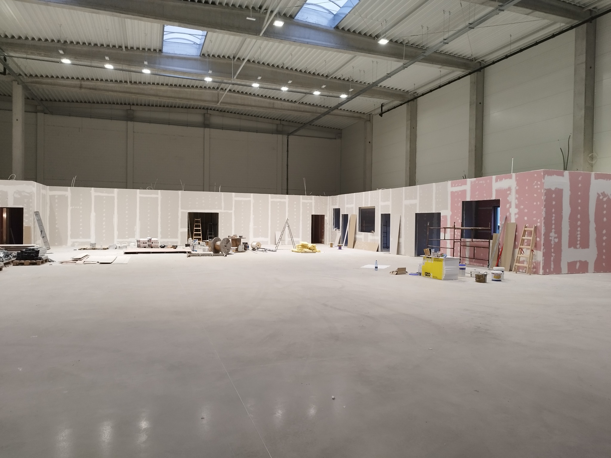 Wnętrze hali produkcyjnej w trakcie adaptacji: widoczne ścianki działowe z płyt gipsowo-kartonowych, otwory drzwiowe, drabiny, materiały budowlane i narzędzia, betonowa posadzka, oświetlenie sufitowe.