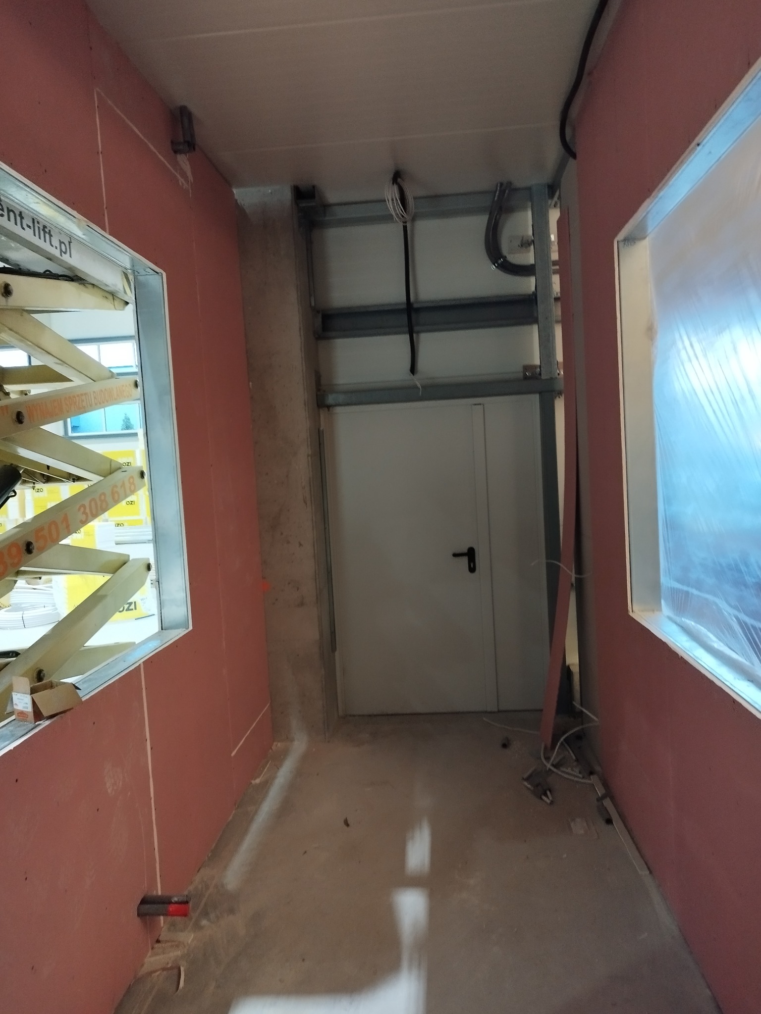 Wnętrze w trakcie prac wykończeniowych: ściany pokryte płytami gipsowo-kartonowymi, okna zabezpieczone folią, widoczna suwnica i drzwi techniczne w głębi.