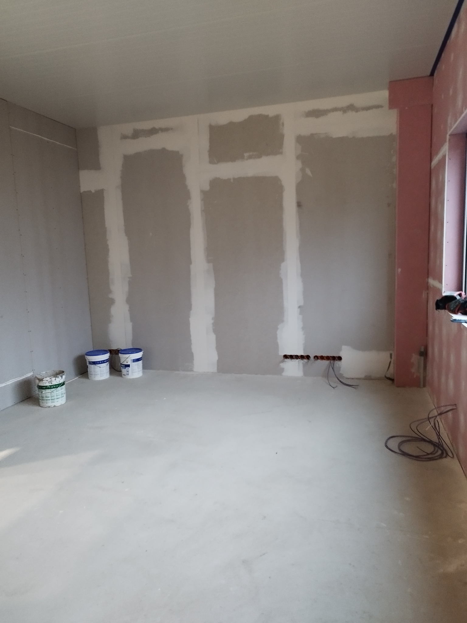 Pokój w trakcie przygotowania do malowania: ściany z nałożonymi pasami gładzi, widoczne puszki z farbą, przewody elektryczne wystające ze ściany i okno z różowym obramowaniem.