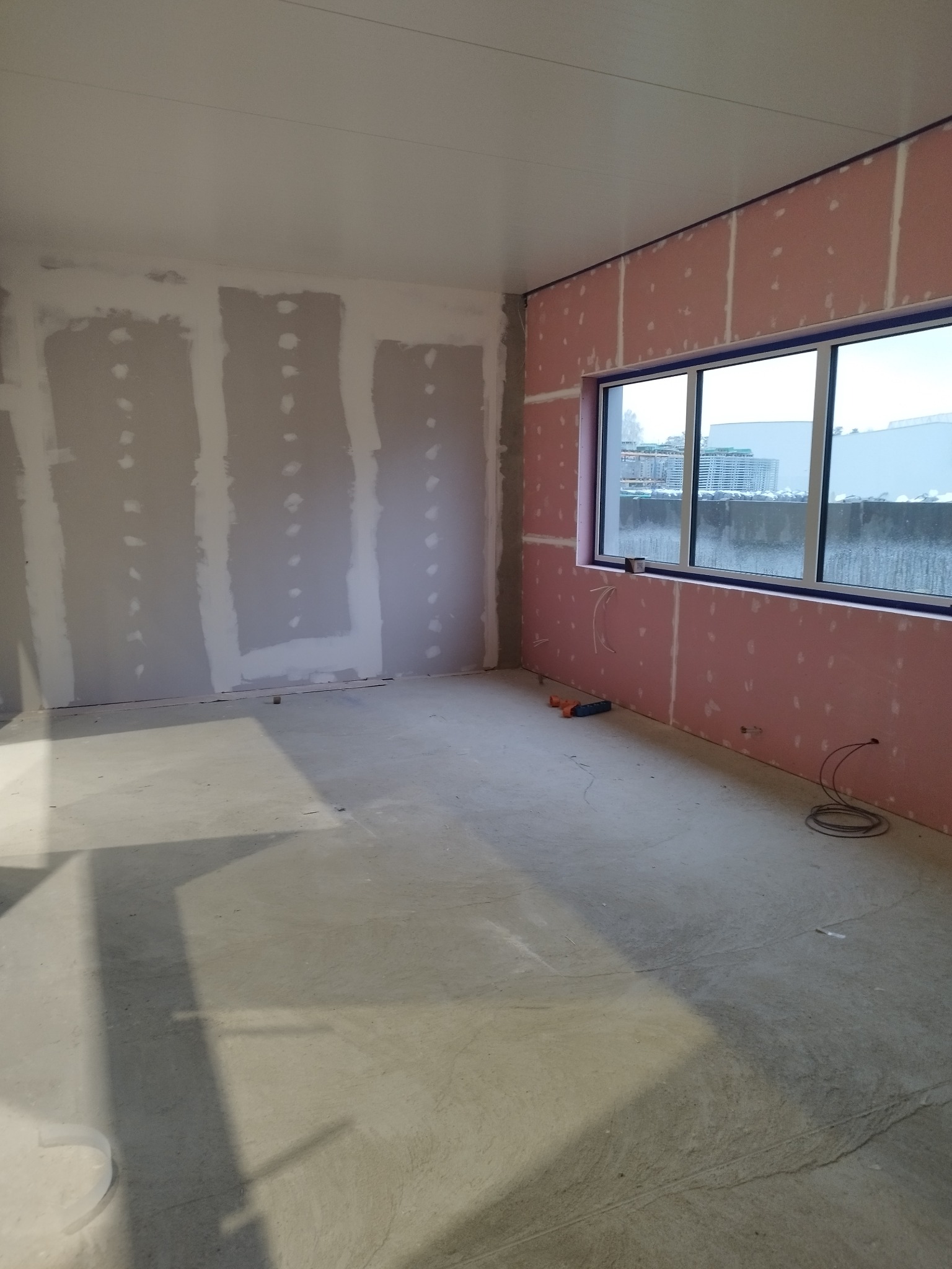 Wnętrze pokoju w trakcie prac wykończeniowych: ściany z płyt gipsowo-kartonowych, częściowo zagruntowane, widoczne narzędzia i przewody elektryczne przy oknie z widokiem na miasto.