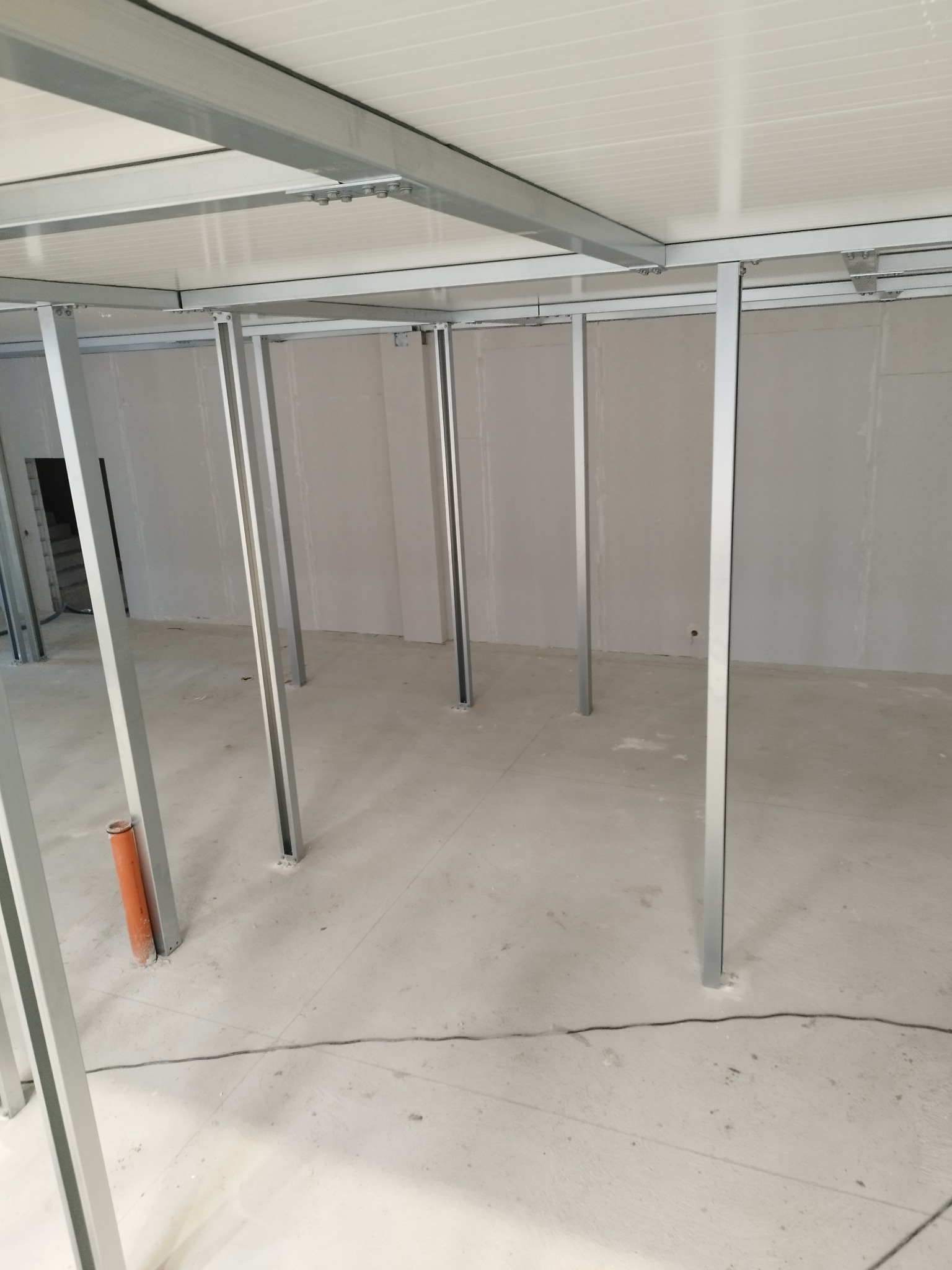 Montaż stelażu sufitu podwieszanego z profili metalowych, widoczne białe panele sufitowe i pomarańczowa rura w rogu pomieszczenia.