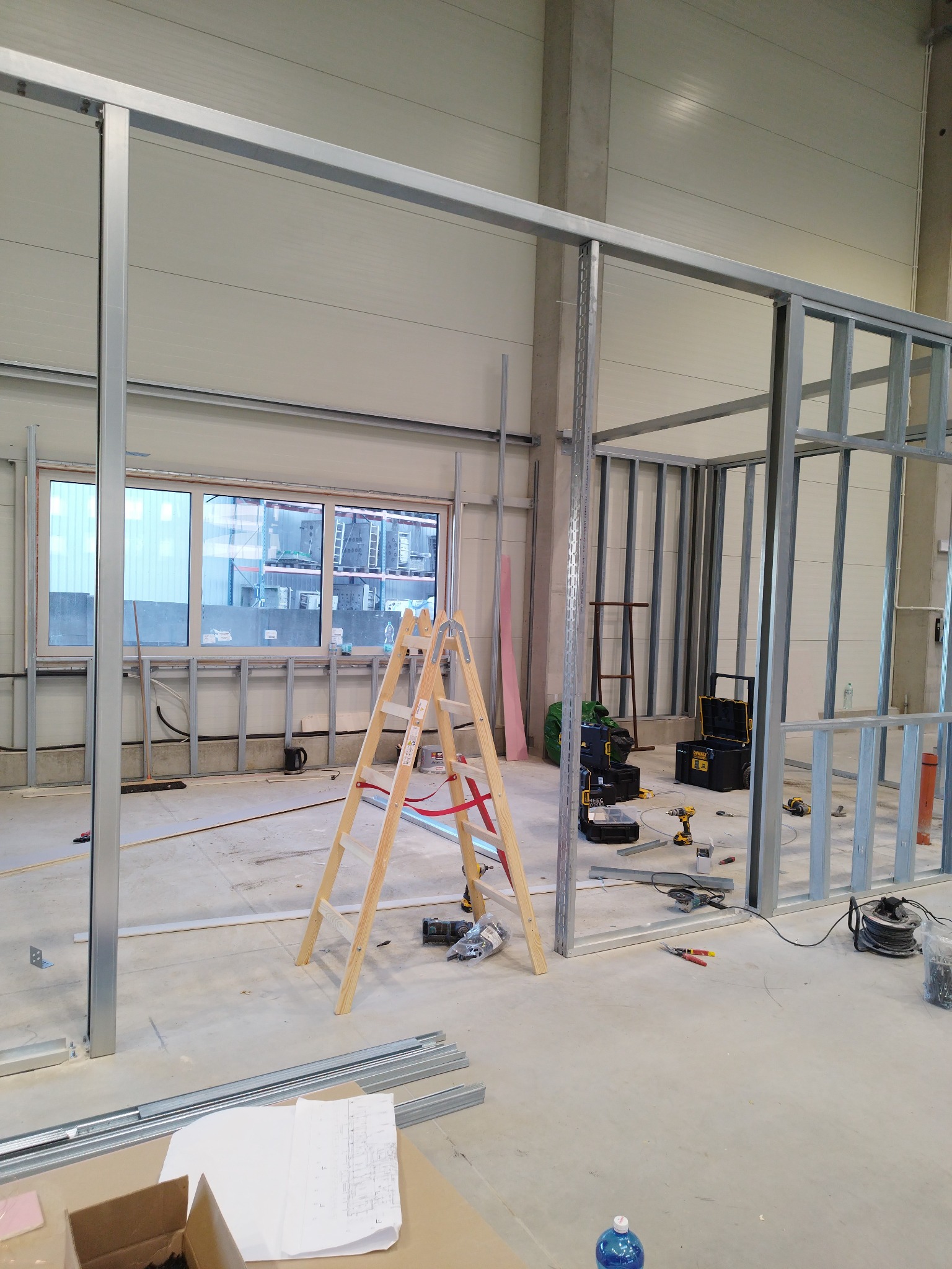 Montaż szkieletu ścian działowych z profili stalowych w surowym wnętrzu, widoczna drabina, narzędzia i plany na podłodze, okna w tle.