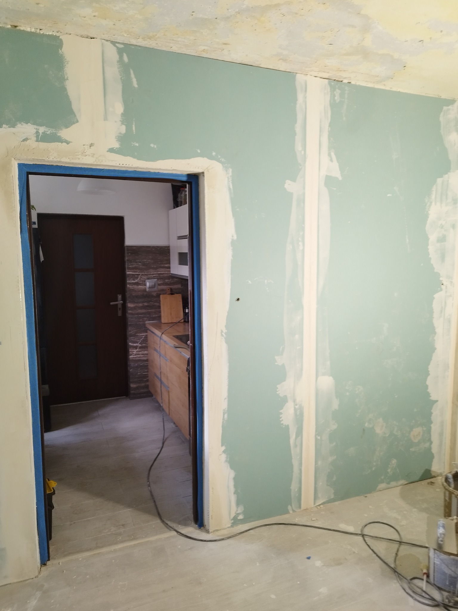 Ściana z płyt gipsowo-kartonowych w trakcie szpachlowania, widoczne łączenia płyt, fragment kuchni w tle przez otwarte drzwi, podłoga w stanie surowym.