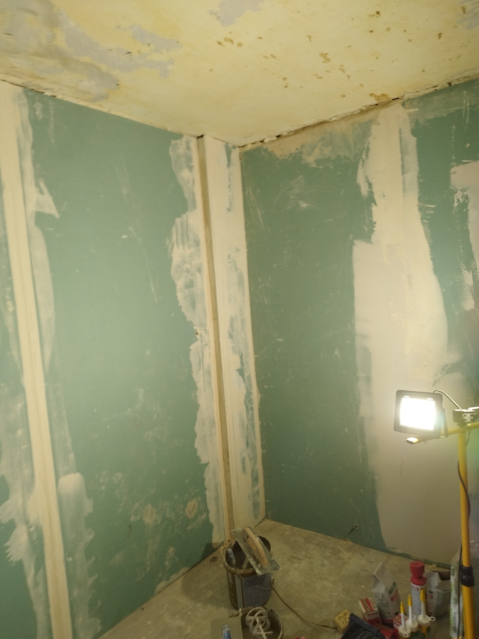 Ściany w trakcie szpachlowania, widoczne ślady po gładzi, narzędzia i oświetlenie budowlane na podłodze, sufit z widocznymi niedoskonałościami.