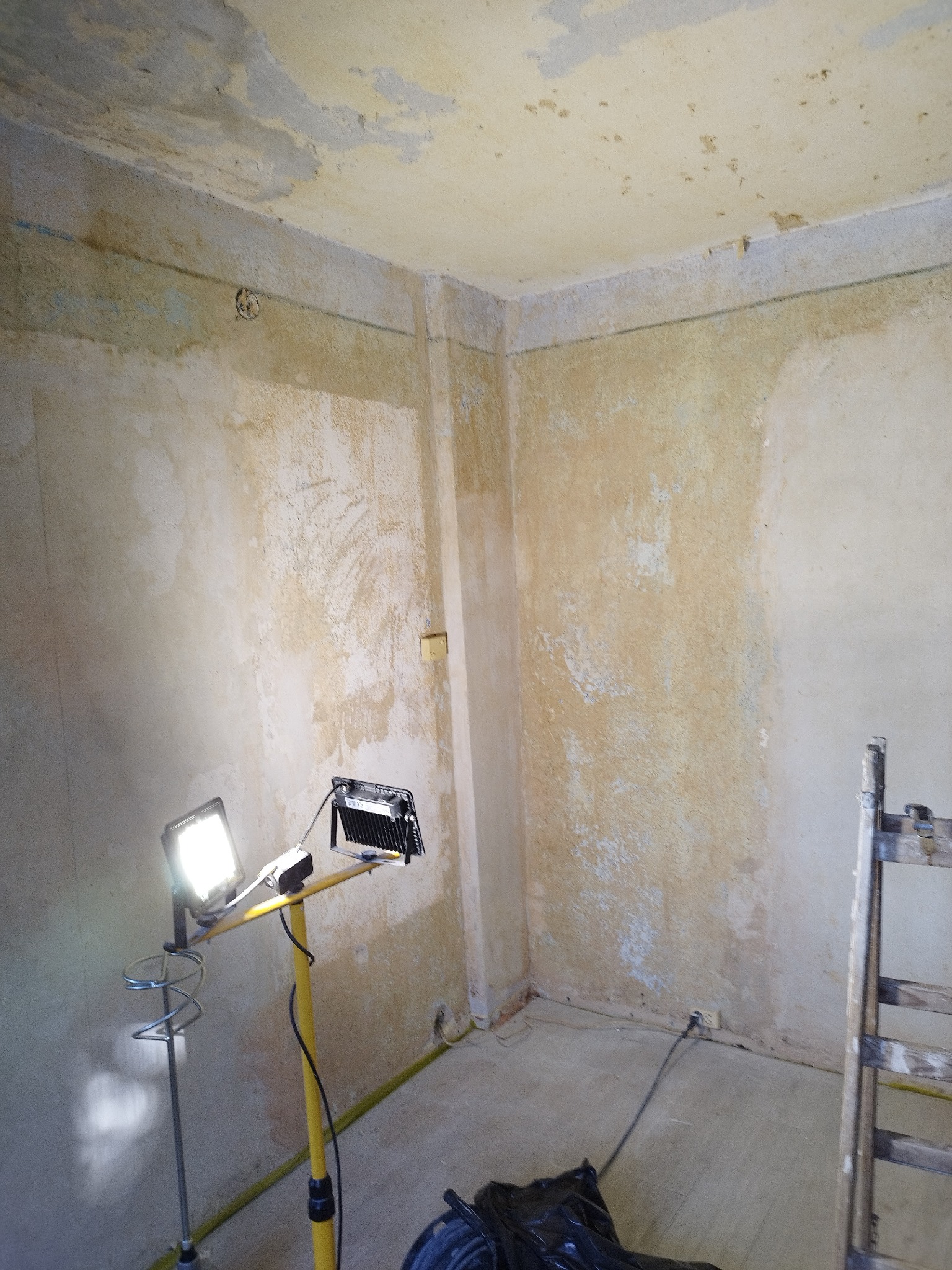 Pokój w trakcie przygotowania do prac wykończeniowych, widoczne ściany ze śladami starej farby i gładzi, oświetlenie robocze na statywie oraz drabina.