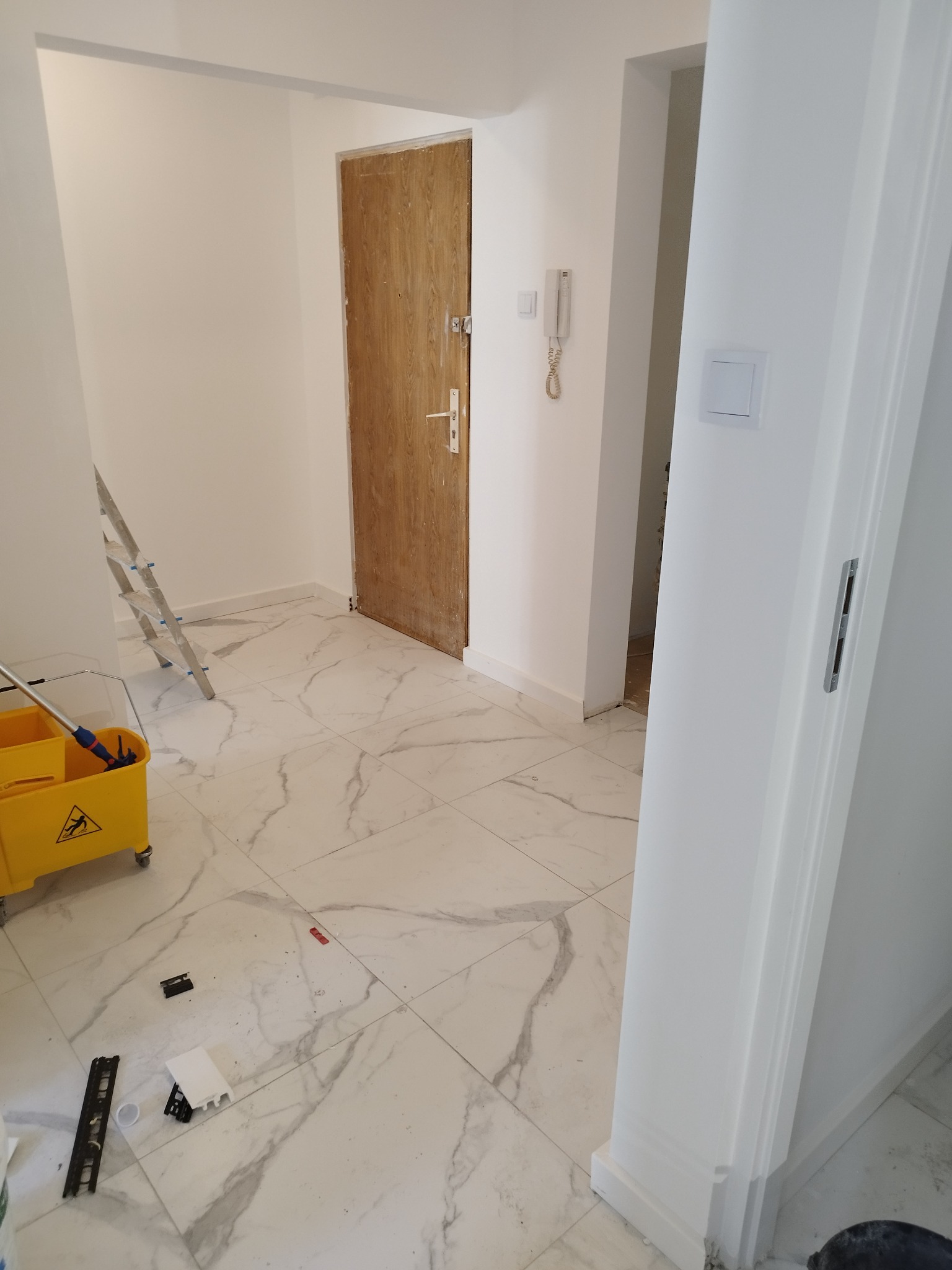 Wnętrze przedpokoju w trakcie malowania: białe ściany, marmurowe płytki podłogowe, żółty wózek z mopem, drabina oparta o ścianę, drzwi wejściowe w tle.