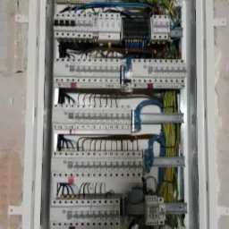 Rozdzielnica elektryczna z licznymi zabezpieczeniami i okablowaniem, zamontowana w surowej ścianie z widocznymi śladami po tynku.