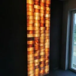 Świecący panel dekoracyjny z podświetlonych, nieregularnych blok&oacute;w w odcieniach pomarańczy i brązu, zamontowany na ciemnej ścianie w nowoczesnym wnętrzu z oknem.