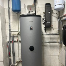 Dolnośląskie Centrum Instalacji - Instalacja kotła grzewczego Hewalex z osprzętem w kotłowni we Wrocławiu. Widoczne rury, zbiorniki wyrównawcze i armatura. Białe płytki na ścianach i podłodze.