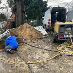 Dolnośląskie Centrum Instalacji - Pracownik w niebieskiej kurtce kuca przy wykopie obok hałdy ziemi. W tle biały van i żółty kompresor na przyczepce. Prace ziemne w błotnistym terenie.