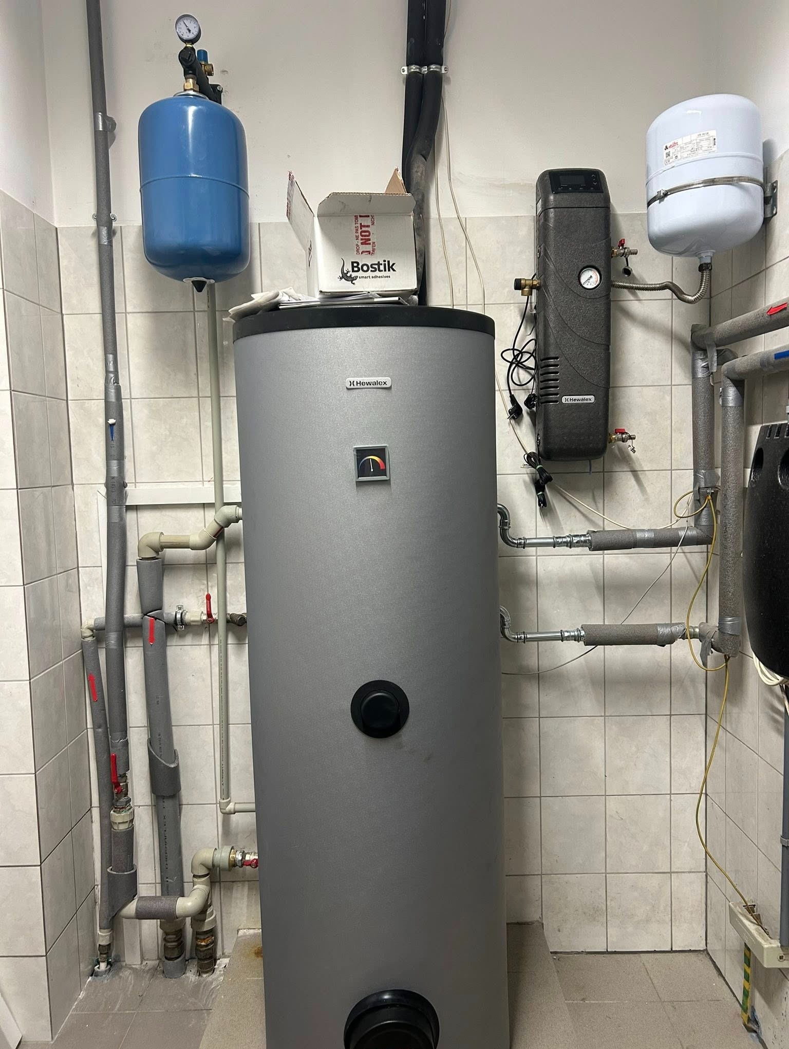 Instalacja kotła grzewczego Hewalex z osprzętem w kotłowni we Wrocławiu. Widoczne rury, zbiorniki wyrównawcze i armatura. Białe płytki na ścianach i podłodze.