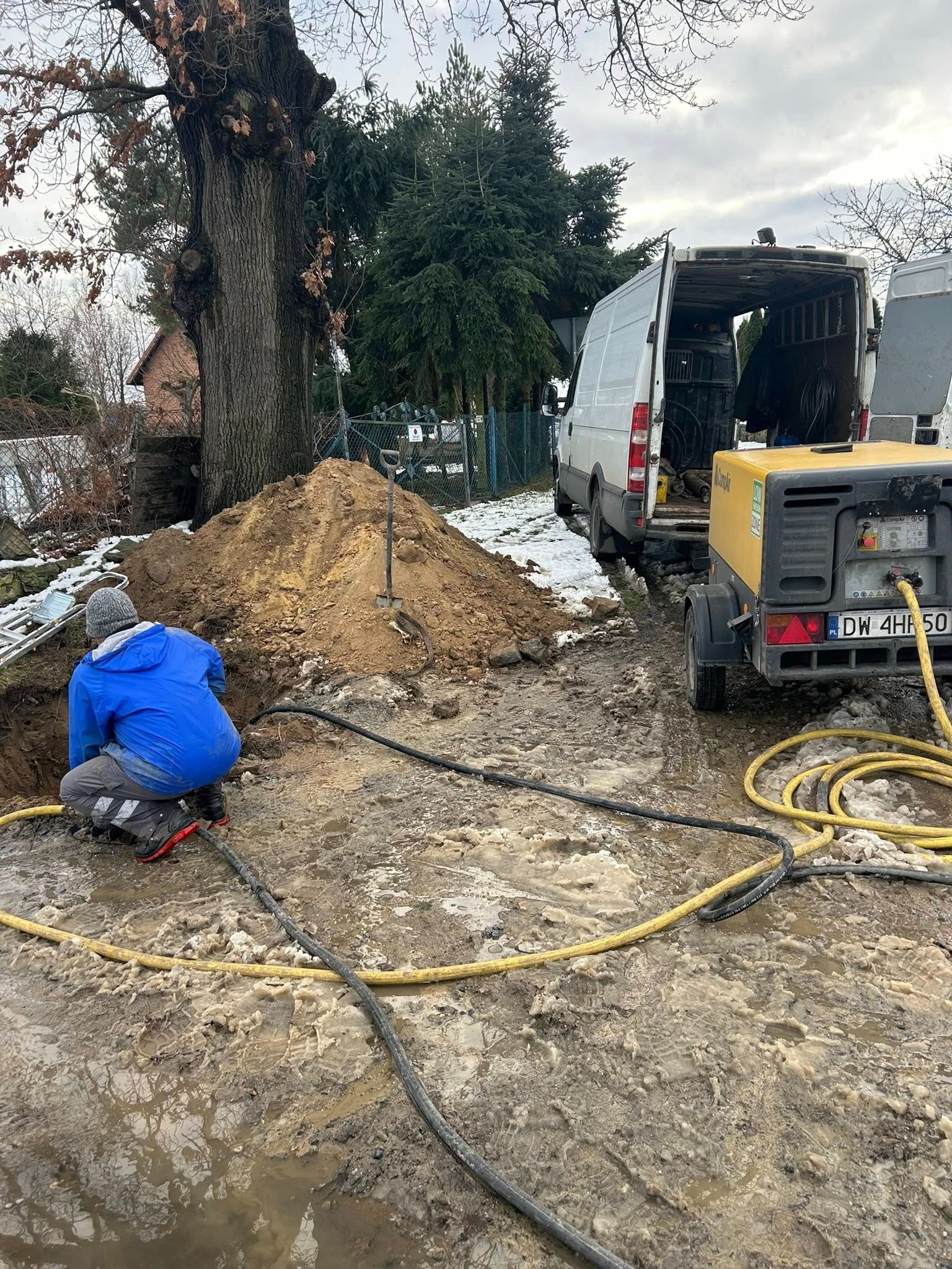 Pracownik w niebieskiej kurtce kuca przy wykopie obok hałdy ziemi. W tle biały van i żółty kompresor na przyczepce. Prace ziemne w błotnistym terenie.