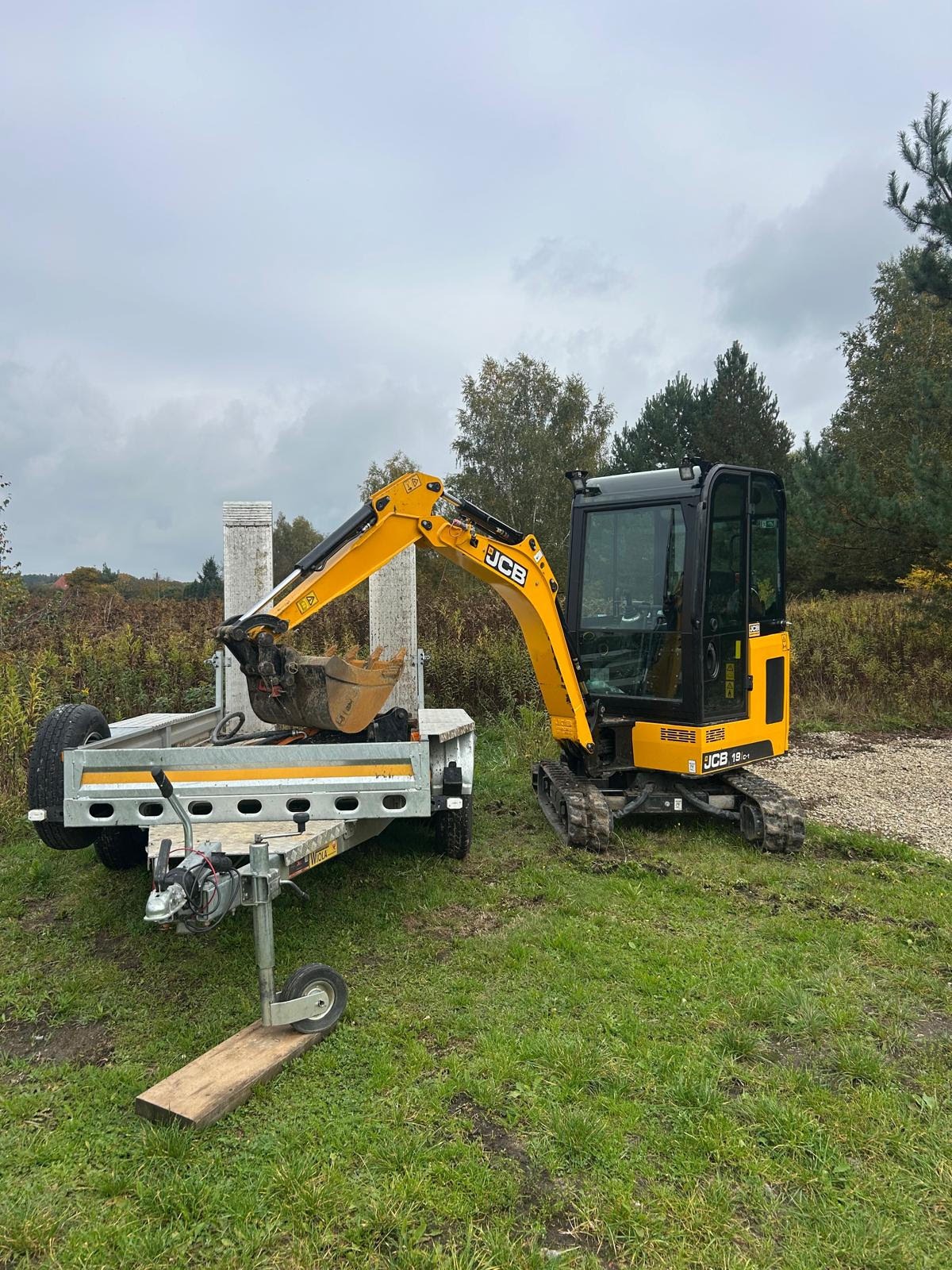 Żółta mini koparka JCB 19C-1 na zielonej trawie obok przyczepy z betonowymi słupkami w tle, pochmurne niebo.