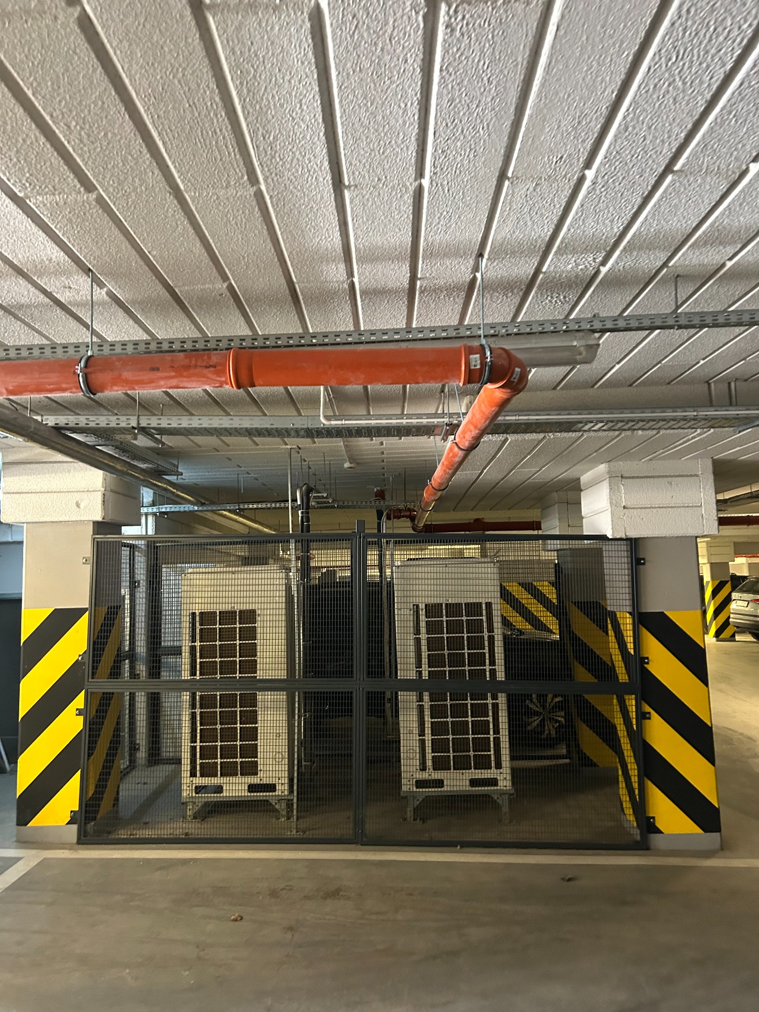 Dwa klimatyzatory za metalową siatką w garażu podziemnym, widoczne pomarańczowe rury wentylacyjne pod sufitem. Słupy oznaczone pasami ostrzegawczymi.