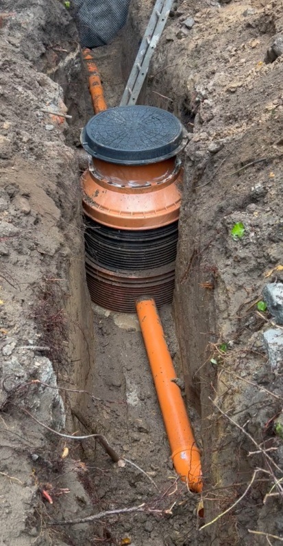 Studzienka kanalizacyjna z rurami w wykopie ziemnym, widok z góry. Elementy instalacji kanalizacyjnej w trakcie montażu, widoczne korzenie i drabina oparta o wykop.