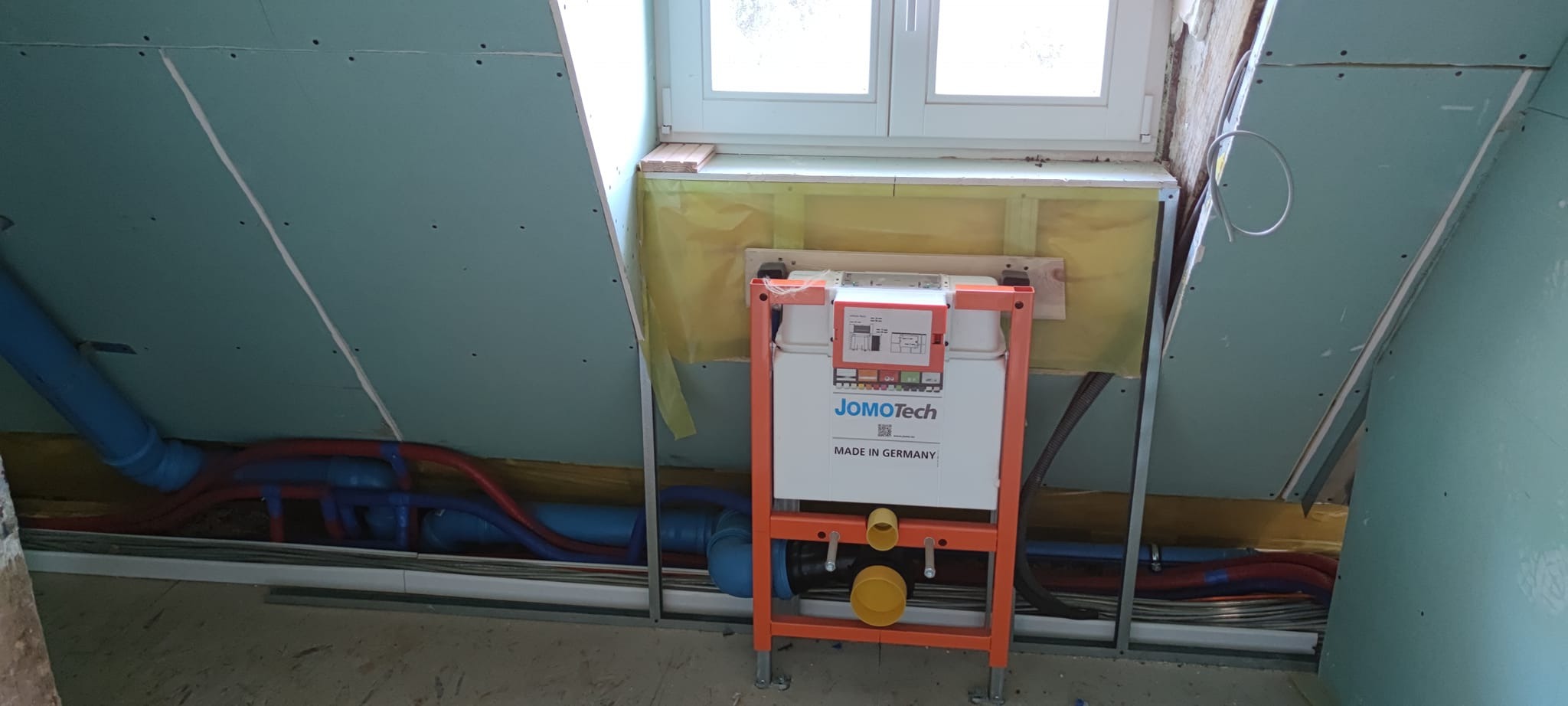 Montaż stelaża podtynkowego JomoTech w łazience na poddaszu, widoczne rury instalacyjne i płyty gipsowo-kartonowe. Prace instalacyjne w toku.