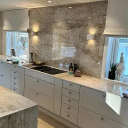 Nowoczesna kuchnia z marmurową ścianą, białymi szafkami i blatem oraz oświetleniem punktowym i ściennym.