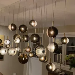 Nowoczesna instalacja oświetleniowa z wieloma szklanymi kulami zawieszonymi na cienkich linkach, z widocznym obrazem na ścianie i oknem w tle.