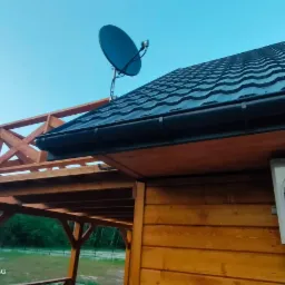 Antena satelitarna zamontowana na dachu drewnianego budynku z klimatyzatorem na ścianie, widok z dołu w słoneczny dzień.