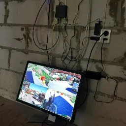 Monitor z podglądem z kamer monitoringu umieszczony na tle ściany z bloczków betonowych z widocznymi kablami i elementami instalacji elektrycznej.