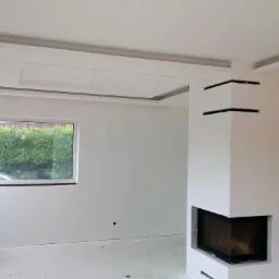 Wnętrze pokoju po remoncie z białymi ścianami, nowoczesnym kominkiem narożnym i ozdobnym sufitem podwieszanym z oświetleniem, widok na zieleń za oknem.