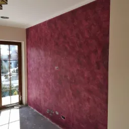 Ściana w pokoju po nałożeniu dekoracyjnej, bordowo-fioletowej struktury, widoczne puszki elektryczne bez osprzętu, okno z widokiem na zieleń.