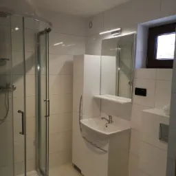 Jasna łazienka z kabiną prysznicową z zaokrąglonymi drzwiami, białą szafką z umywalką, lustrem z oświetleniem LED i oknem z ciemną ramą.