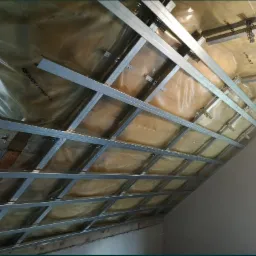 Skośny sufit poddasza w trakcie montażu stelaża z profili stalowych, widoczna izolacja z folii i wełny mineralnej, oświetlenie robocze, drabina w rogu pomieszczenia.