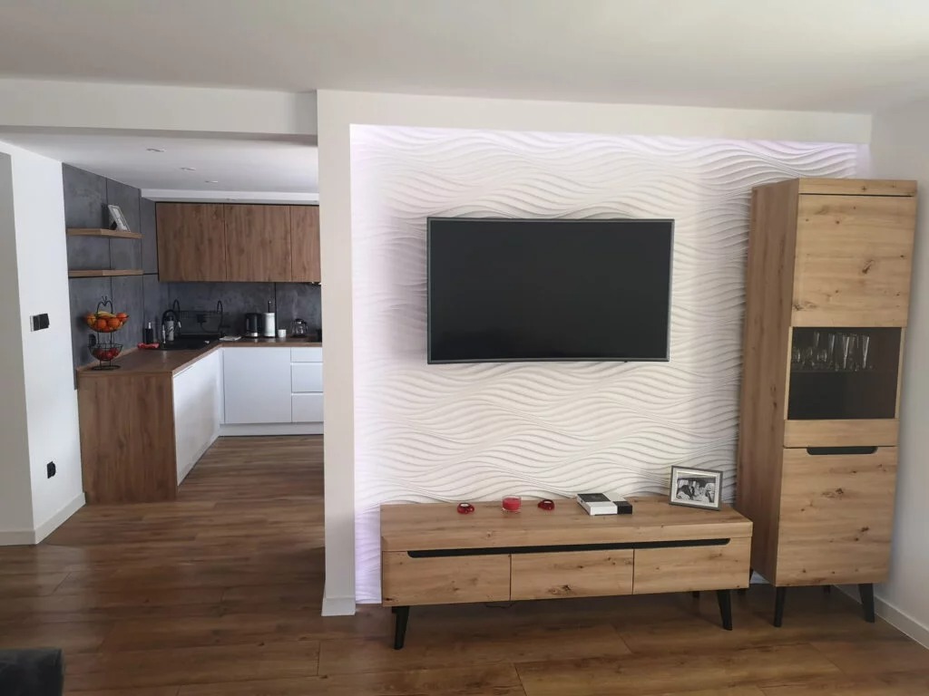 Nowoczesne wnętrze: drewnopodobna podłoga, biała kuchnia widoczna w tle, ściana z falistym wzorem, telewizor, szafka RTV i wysoka witryna w odcieniach drewna.