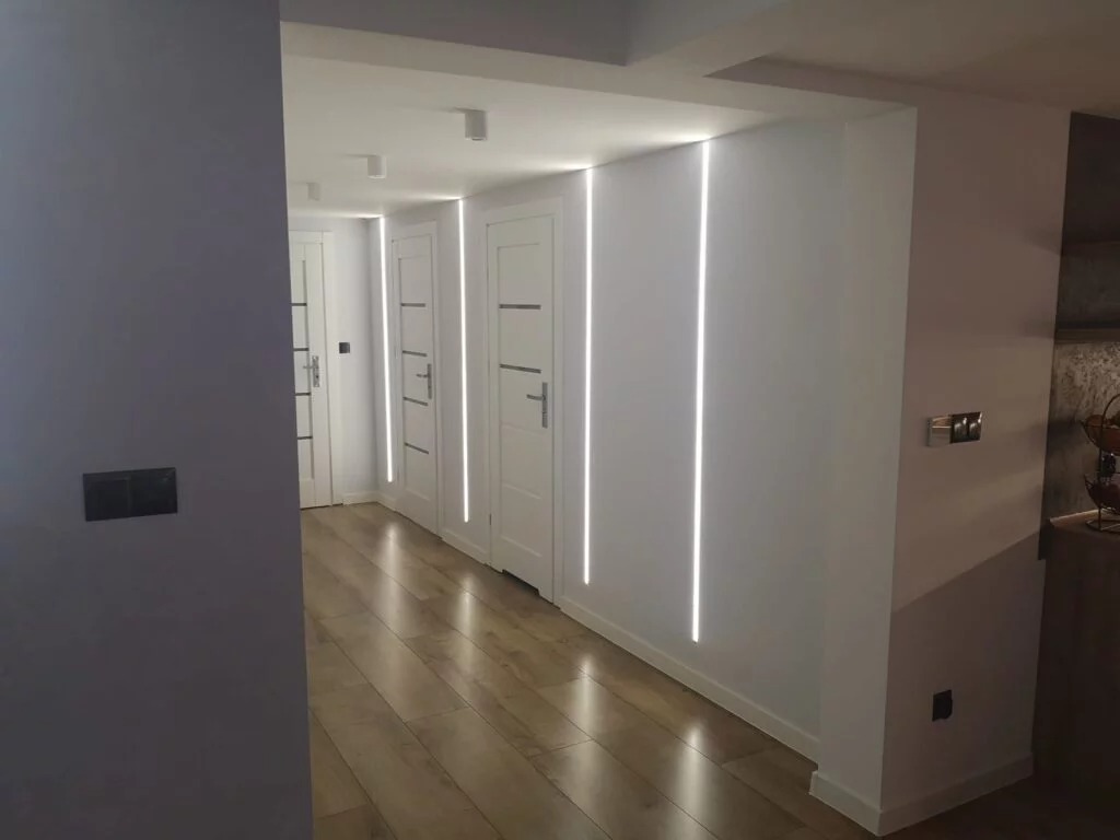 Korytarz z trzema białymi drzwiami, podświetlonymi pionowymi paskami LED, parkiet ułożony w jodełkę, szara ściana po lewej stronie.