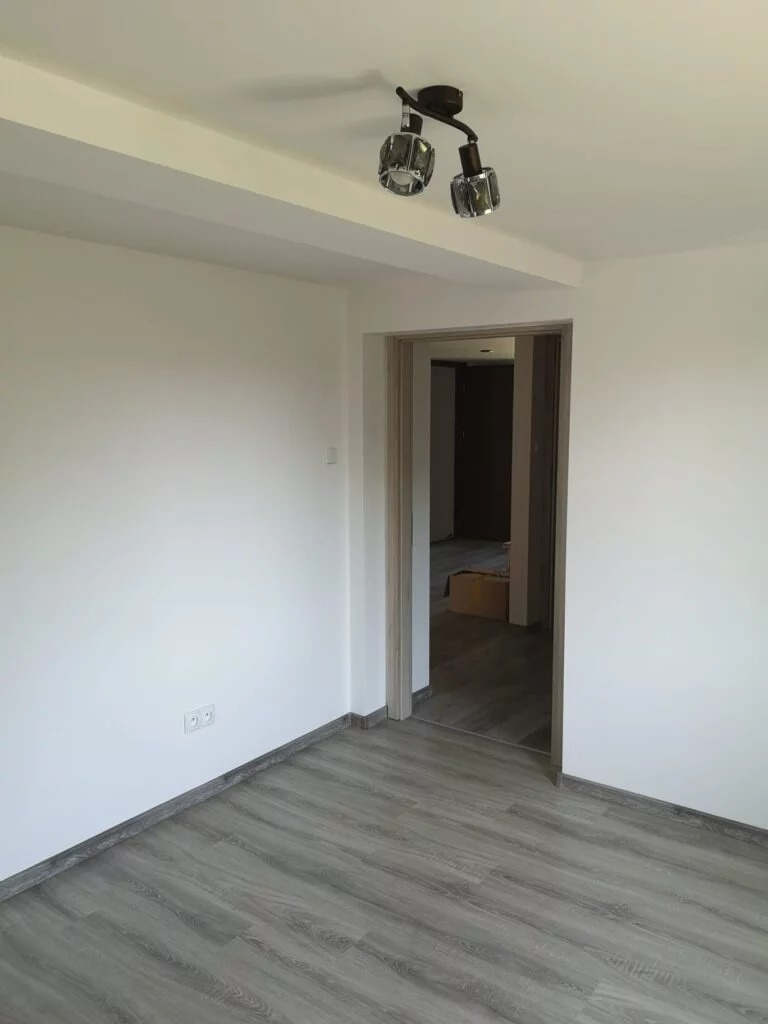 Pusty pokój po remoncie z szarymi panelami podłogowymi, białymi ścianami i nowoczesnym oświetleniem sufitowym z dwoma reflektorami.