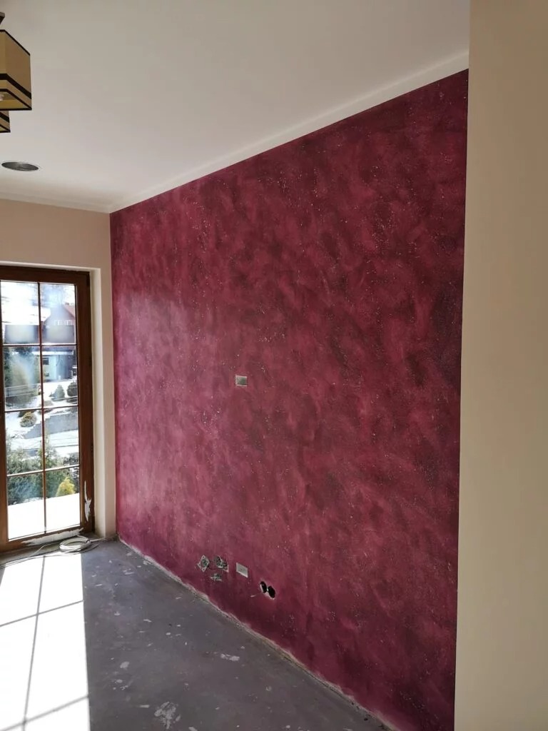 Ściana w pokoju po nałożeniu dekoracyjnej, bordowo-fioletowej struktury, widoczne puszki elektryczne bez osprzętu, okno z widokiem na zieleń.