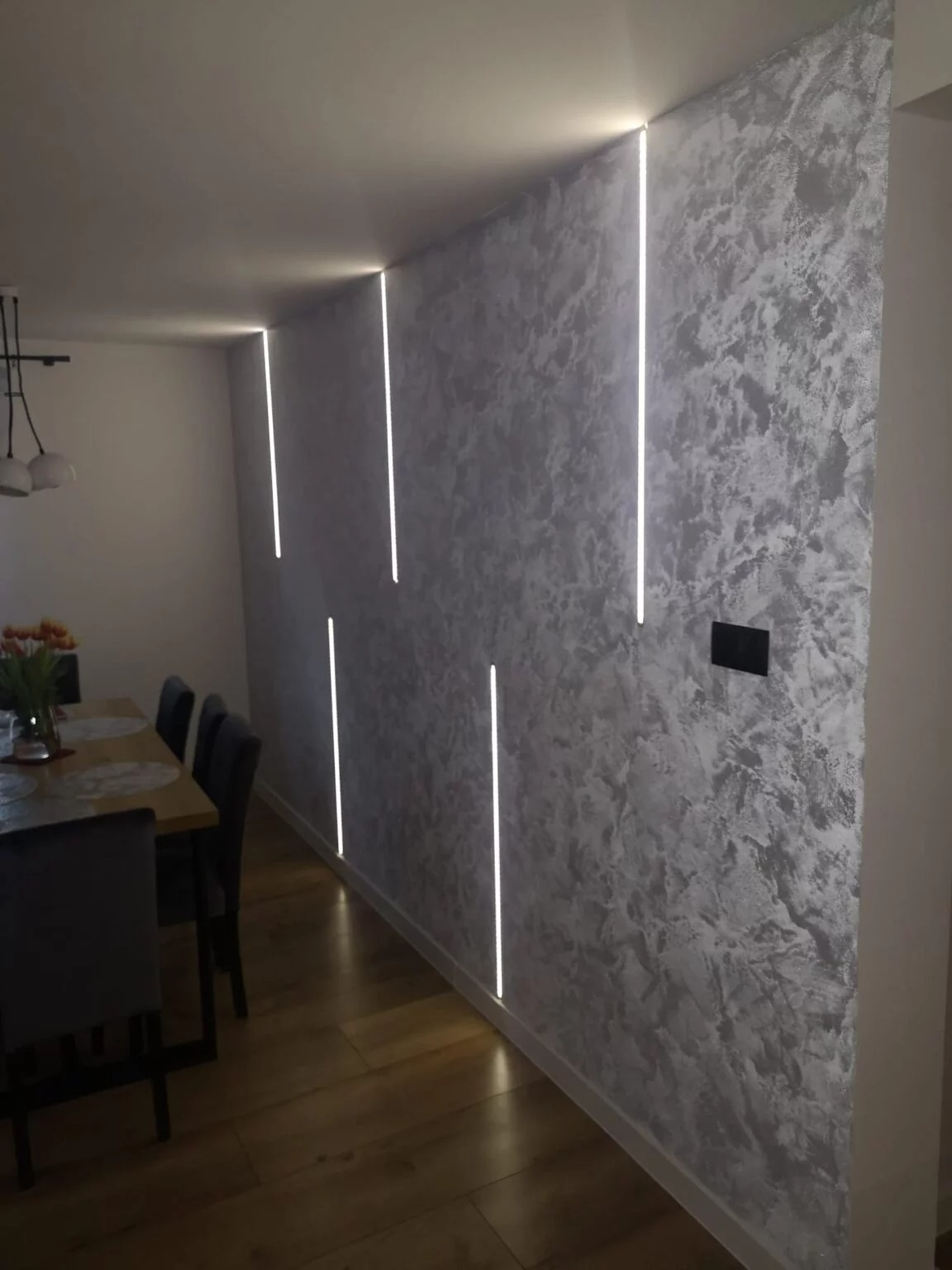 Ściana z dekoracyjną szarą strukturą i pionowymi liniami oświetlenia LED, widoczny fragment podłogi z paneli oraz meble w tle.