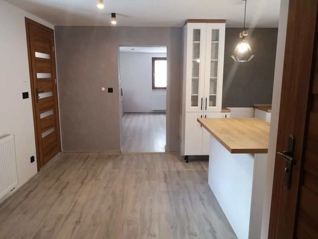 Jasne panele podłogowe w nowocześnie wykończonym wnętrzu z szarą ścianą, drewnianymi drzwiami i białą zabudową kuchenną z drewnianym blatem oraz ozdobną lampą.