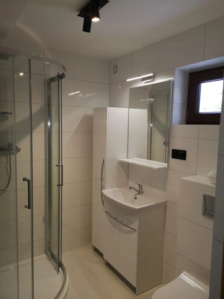 Jasna łazienka z kabiną prysznicową z zaokrąglonymi drzwiami, białą szafką z umywalką, lustrem z oświetleniem LED i oknem z ciemną ramą.