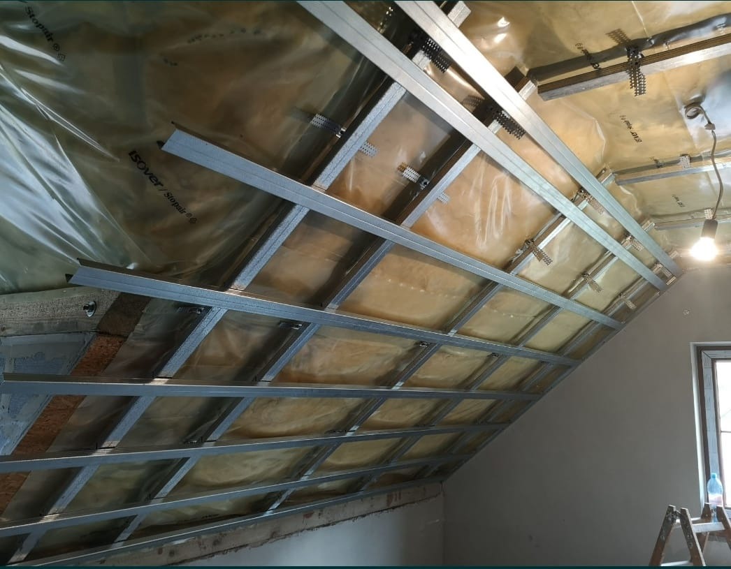 Skośny sufit poddasza w trakcie montażu stelaża z profili stalowych, widoczna izolacja z folii i wełny mineralnej, oświetlenie robocze, drabina w rogu pomieszczenia.