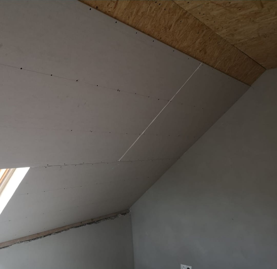 Poddasze z zamontowaną płytą kartonowo-gipsową na skośnym suficie, widoczne wkręty mocujące i łączenie płyt, fragment okna dachowego w lewym dolnym rogu.