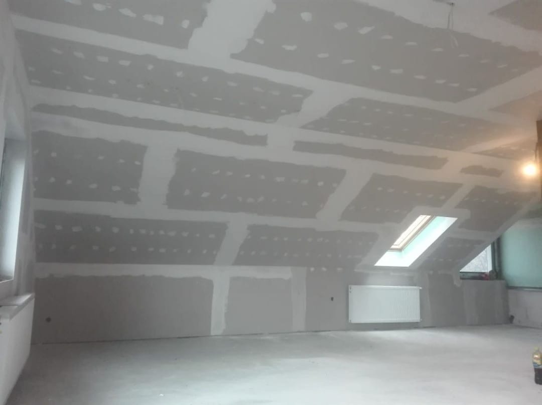 Poddasze z zamontowanymi płytami kartonowo-gipsowymi, widoczne łączenia płyt zaszpachlowane, okno dachowe w skosie, grzejnik na ścianie, surowy beton na podłodze.