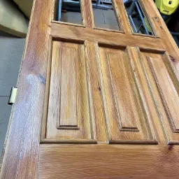 Odrestaurowane, drewniane drzwi z szybami, leżące na boku, widoczne ślady świeżo nałożonej bejcy lub lakieru na części powierzchni, zawias po lewej stronie.