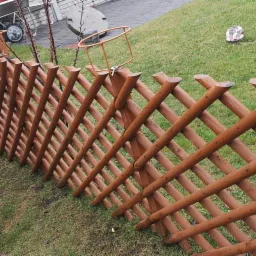 Brązowa, drewniana krata ogrodowa, ułożona na trawie, z widocznymi metalowymi uchwytami na doniczki i fragmentem zieleni w tle.