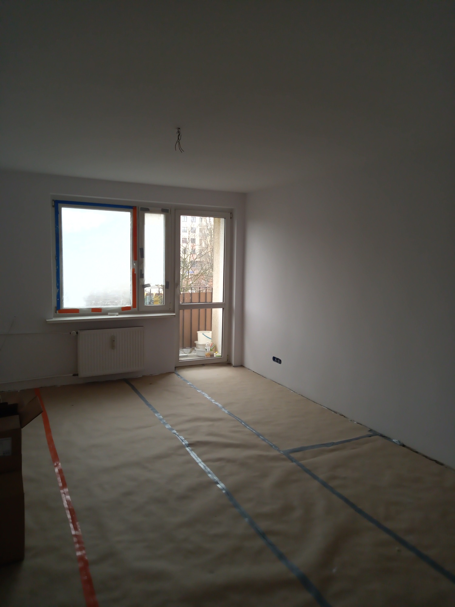 Pokój w trakcie przygotowania do prac wykończeniowych: zabezpieczona podłoga brązowym papierem i taśmą, okna z częściowo oklejonymi ramami, świeżo pomalowane ściany na biało, widoczne kable...