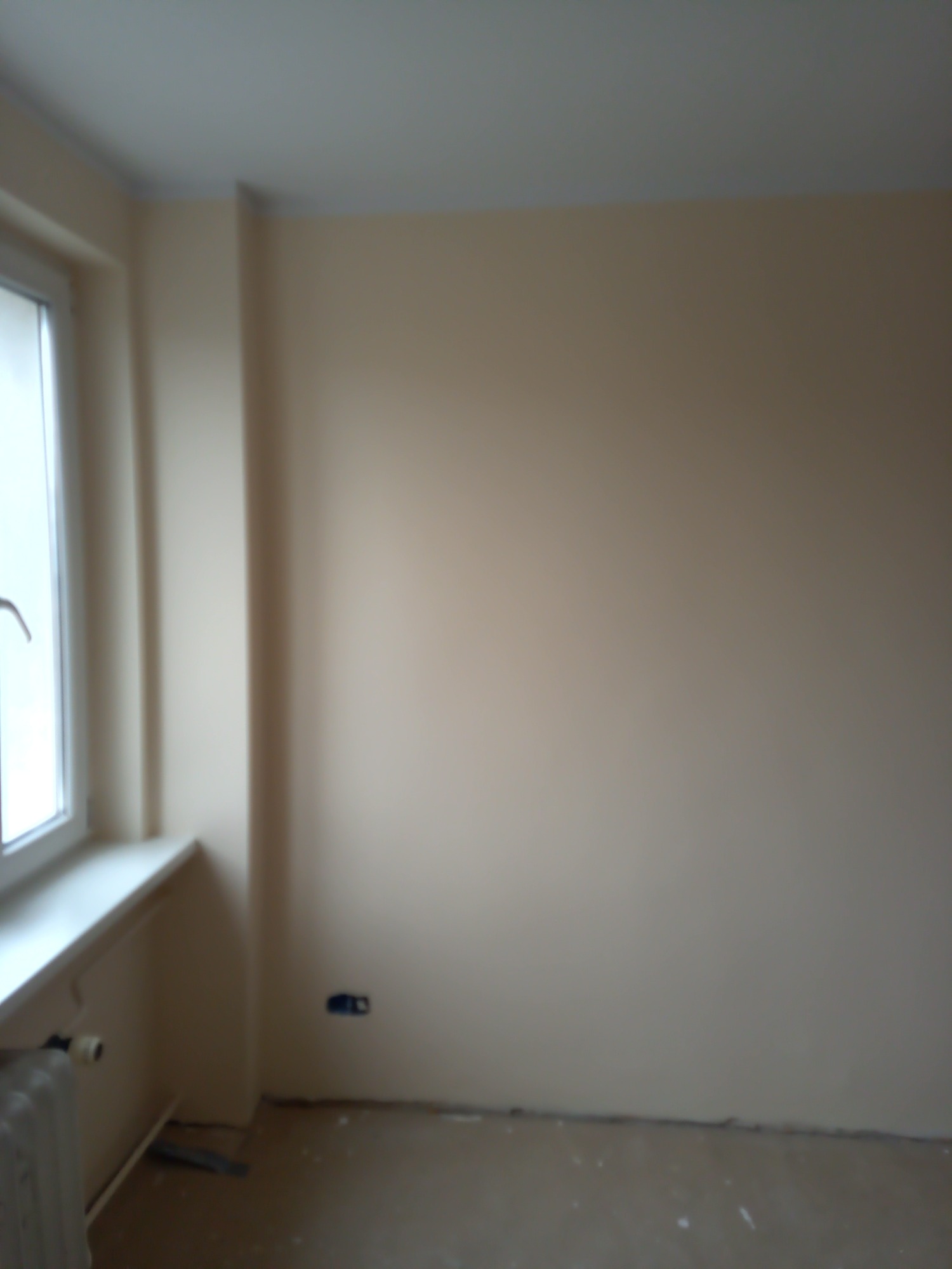 Świeżo pomalowane wnętrze pokoju w beżowym odcieniu, widoczne gniazdko elektryczne na ścianie, grzejnik pod oknem i przygotowane podłoże.