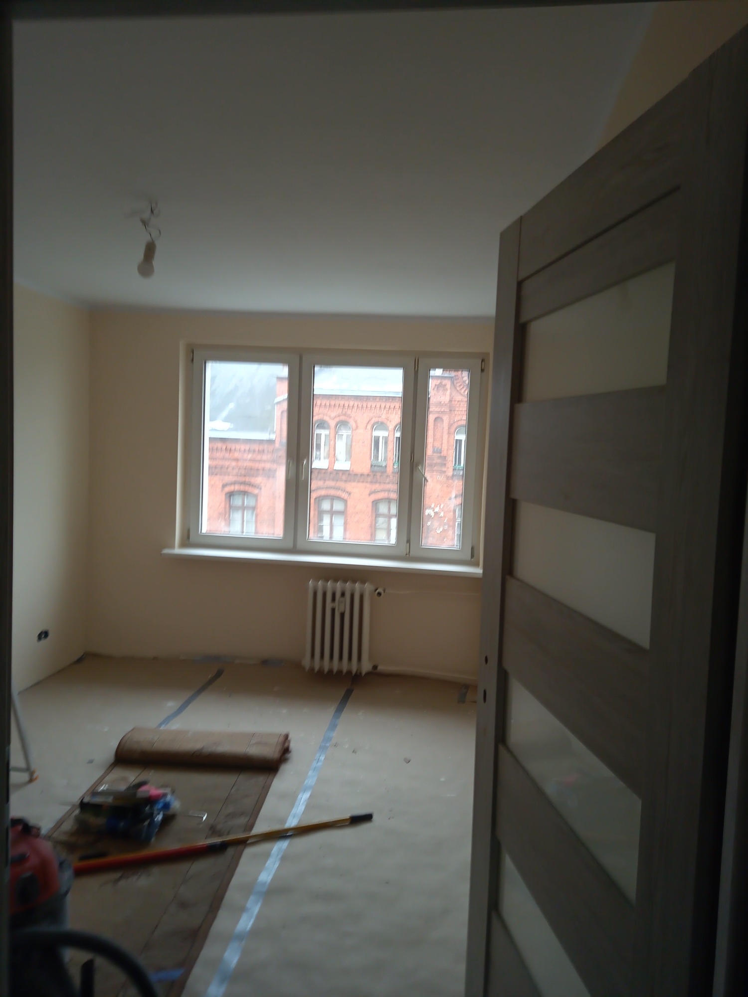 Pokój w trakcie przygotowań do malowania: podłoga zabezpieczona kartonem i taśmą, widoczne narzędzia malarskie, budynek z czerwonej cegły za oknem.