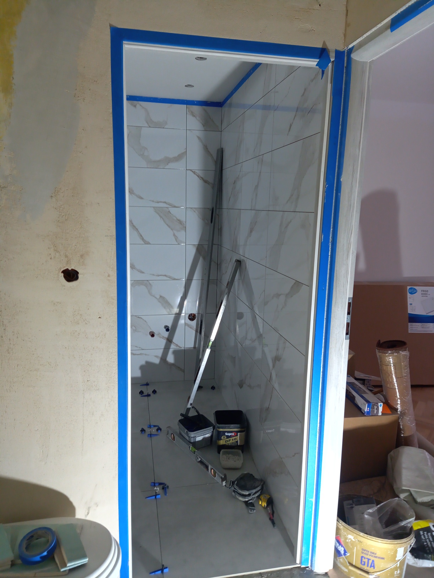 Wnętrze łazienki w trakcie remontu: białe płytki imitujące marmur na ścianach, poziomica oparta o ścianę, niebieska taśma malarska zabezpieczająca framugę, narzędzia i materiały budowlane...