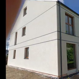 Dwupiętrowy dom z jasną elewacją i brązowymi ramami okiennymi, widoczne szare poziome pasy dekoracyjne, rynna i fragment gruntu przy fundamencie.