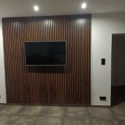 Nowoczesny salon z dekoracyjną ścianą z drewnianych listew, na której zawieszony jest telewizor. Szare płytki na podłodze, białe ściany z gniazdkami elektrycznymi. Oświetlenie sufitowe punktowe.