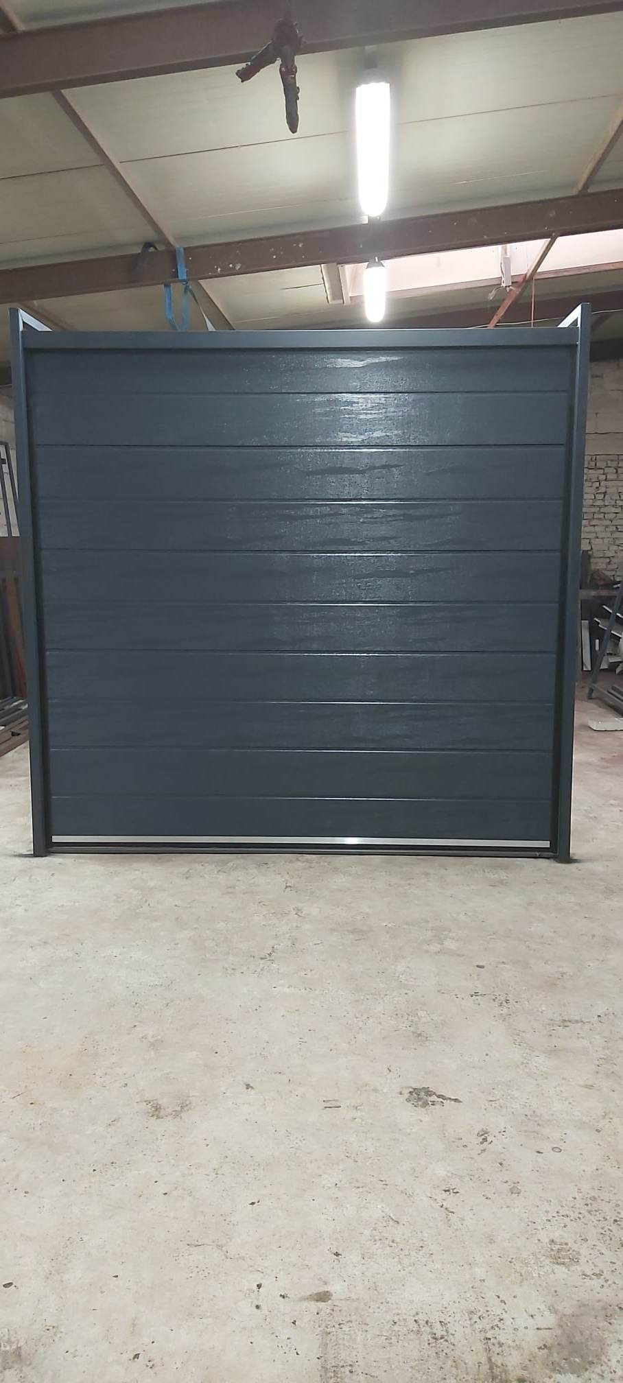 Szara, segmentowa brama garażowa w hali produkcyjnej, z widocznymi poziomymi panelami i metalową ramą, oświetlona lampą jarzeniową.