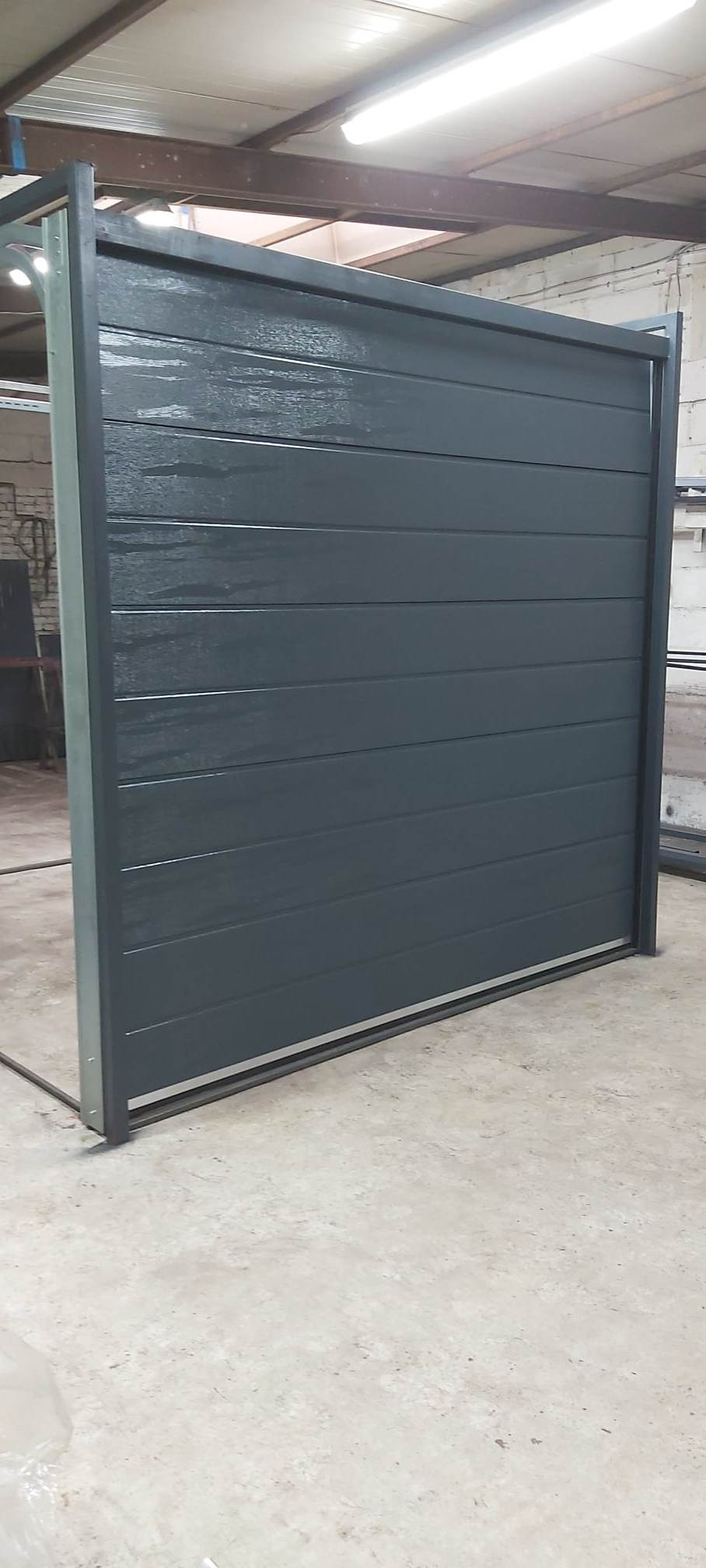 Szara segmentowa brama garażowa z metalową ramą, widoczna w hali produkcyjnej, z połyskującą powierzchnią i poziomymi panelami.