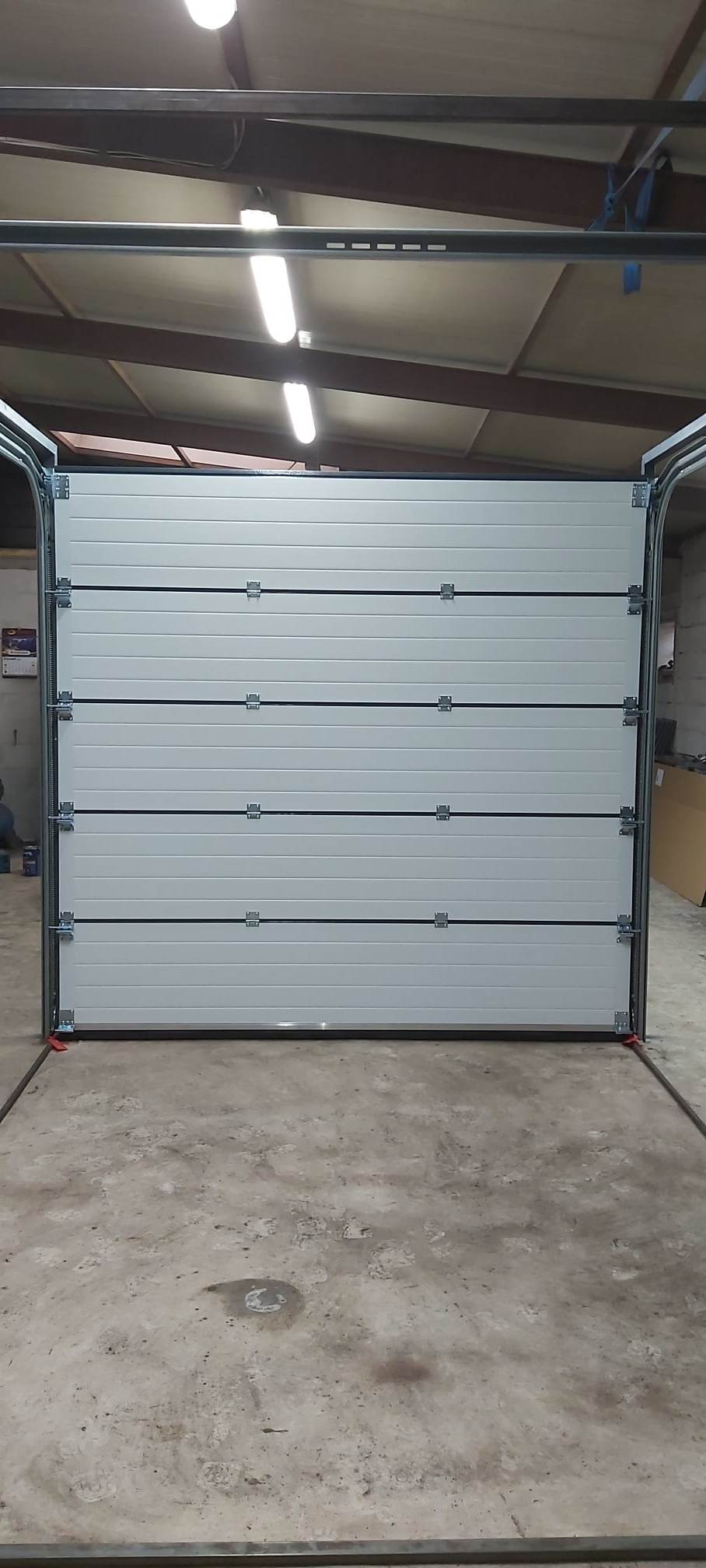 Zamknięta segmentowa brama garażowa w kolorze jasnoszarym, widoczne metalowe prowadnice i elementy mocujące, szara betonowa posadzka w garażu.